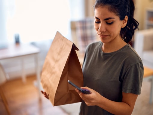 Smiling woman holding a paper bag and checking her food delivery order on a smartphone