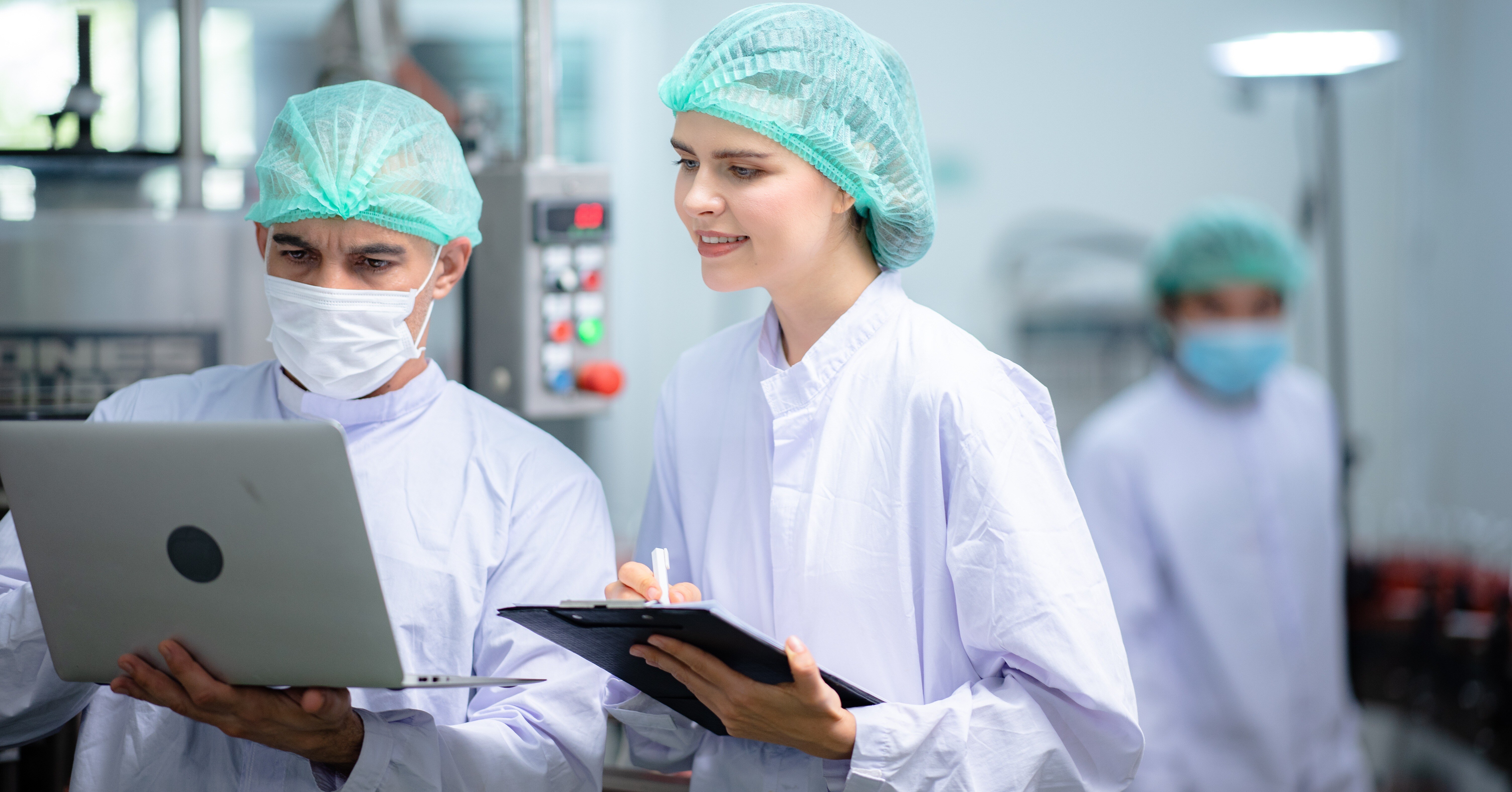 Two food safety inspectors in protective gear review compliance data on a laptop and clipboard inside a food manufacturing facility.